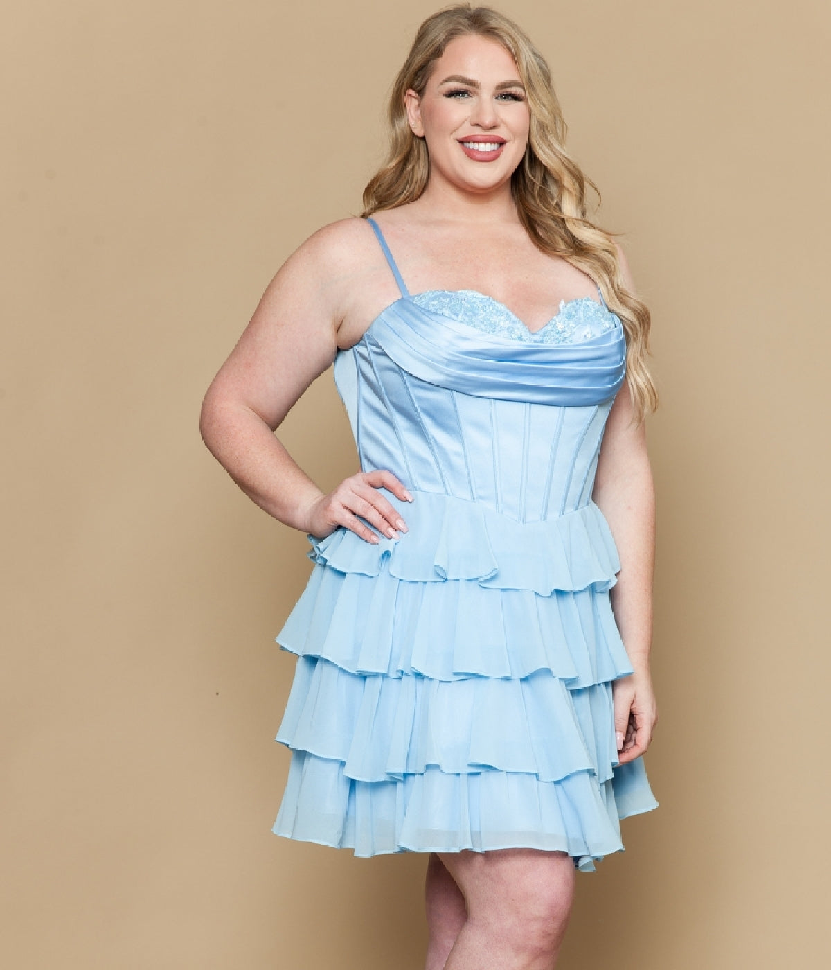 A woman smiles in the Poly USA W1196 Short Plus Size Homecoming Dress, featuring blue tiered ruffles, spaghetti straps, and a satin corset bodice, set against a beige background.